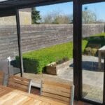 A contemporary round planter filled with dark mulch stands in a small courtyard with overgrown green grass and a white wall featuring a window