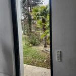 A view through a glass door, suggesting potential windows and door repair needs, into a small, enclosed courtyard with overgrown grass, a potted plant