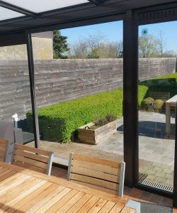 A contemporary round planter filled with dark mulch stands in a small courtyard with overgrown green grass and a white wall featuring a window